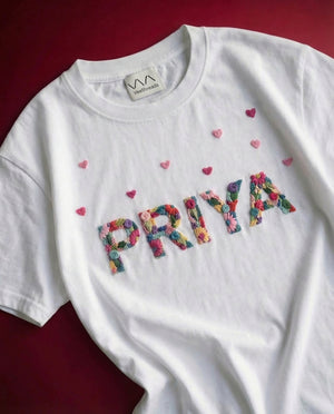 Custom name embroidered t-shirt with hearts — white cotton tee with colourful 'PRIYA' hand-embroidered with small hearts on red background