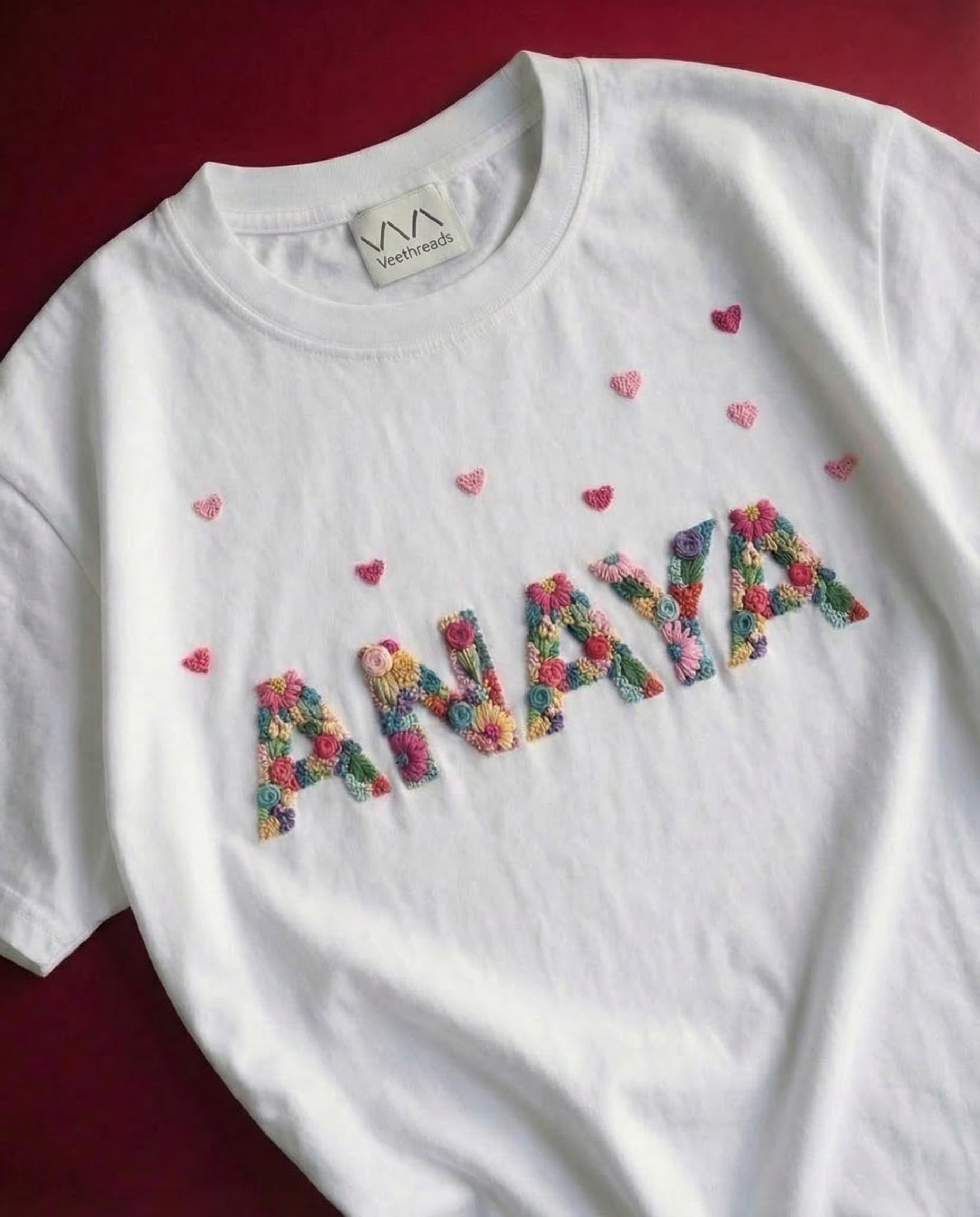 Simple name embroidery designs hearts — white tee with colourful 'ANAYA' hand-embroidered letters and small hearts on red background