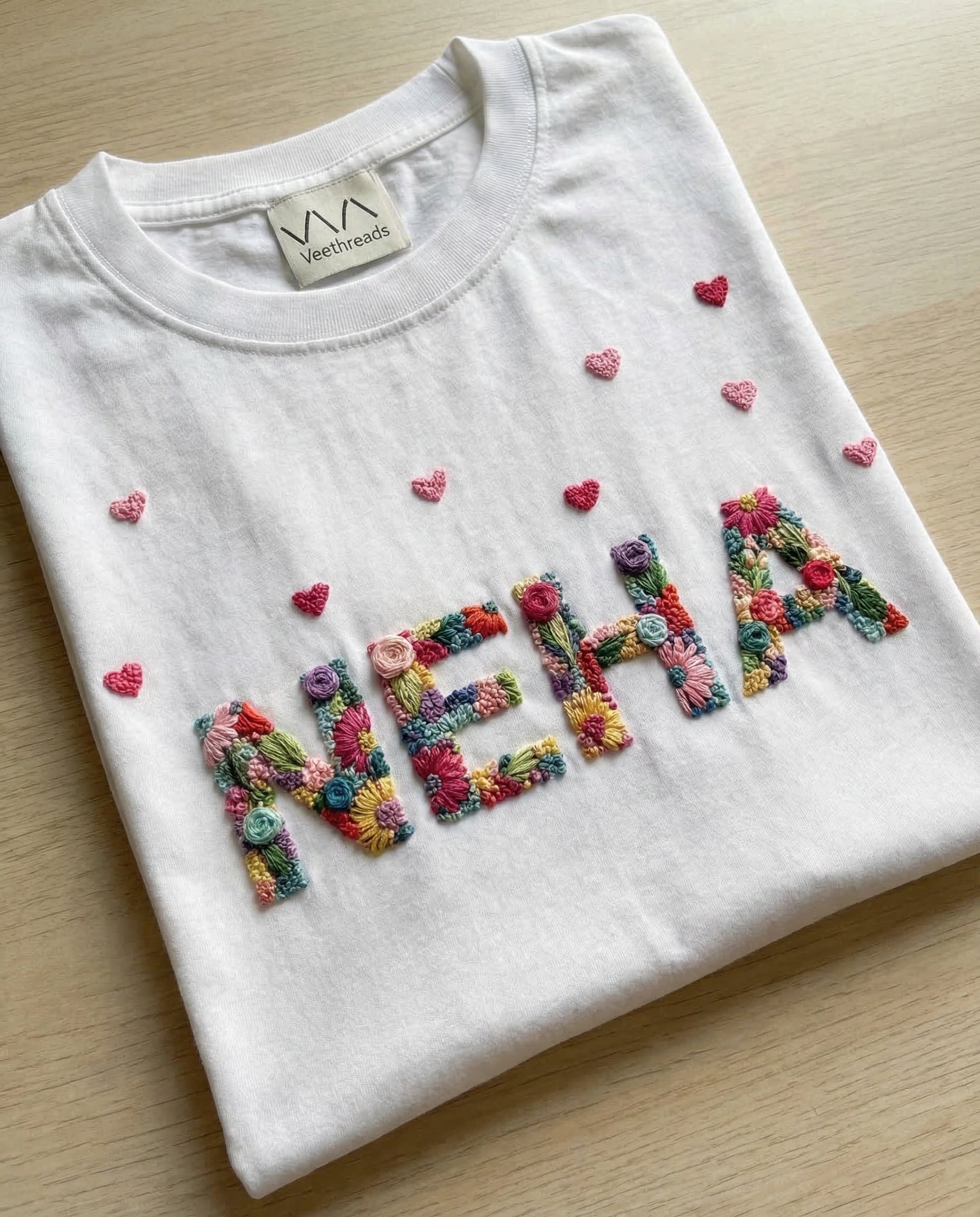 White t-shirt with colorful embroidered letters spelling 'NEHA' on a wooden surface