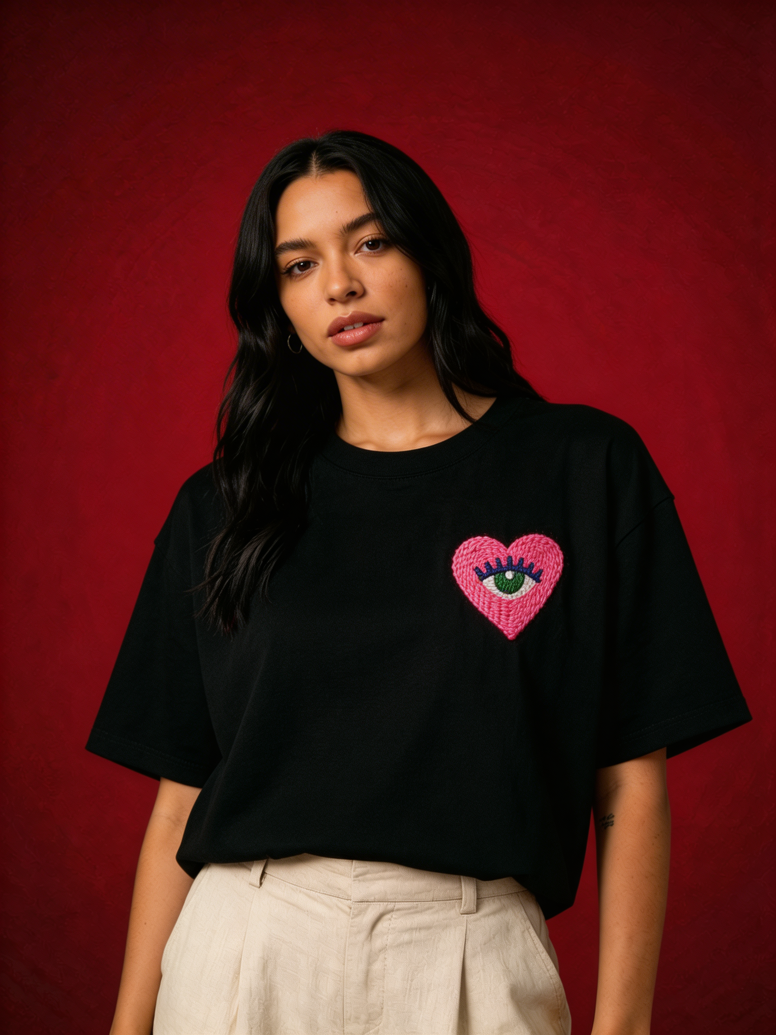 Hand-embroidered pink heart with a green eye on a black cotton tee — Love Eye design by Vee Threads, raised real-thread embroidery.