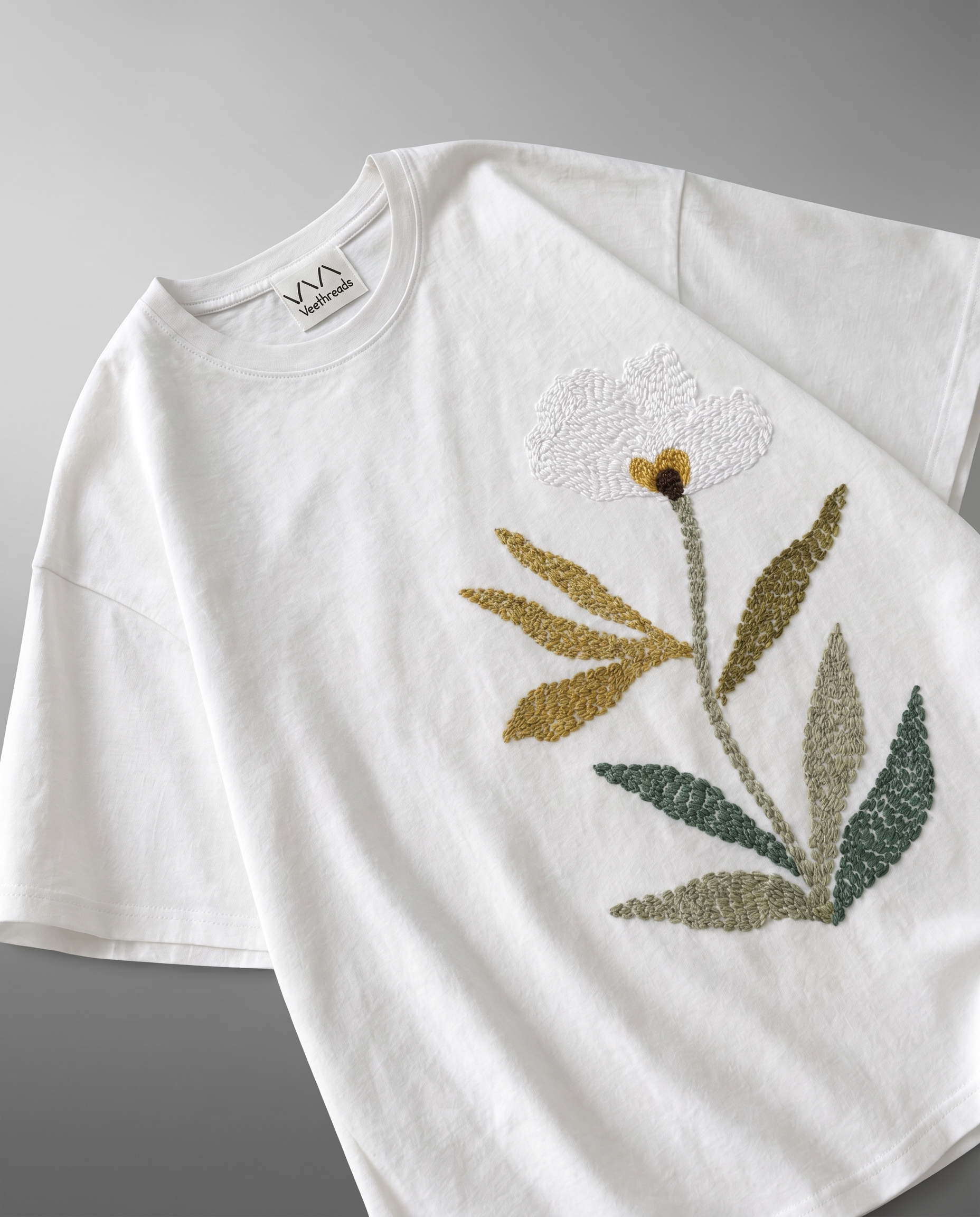 
Hand embroidered botanical bloom detail on white cotton tee — satin stitch white petals, mustard gold stem, olive and sage green leaves by Veethreads