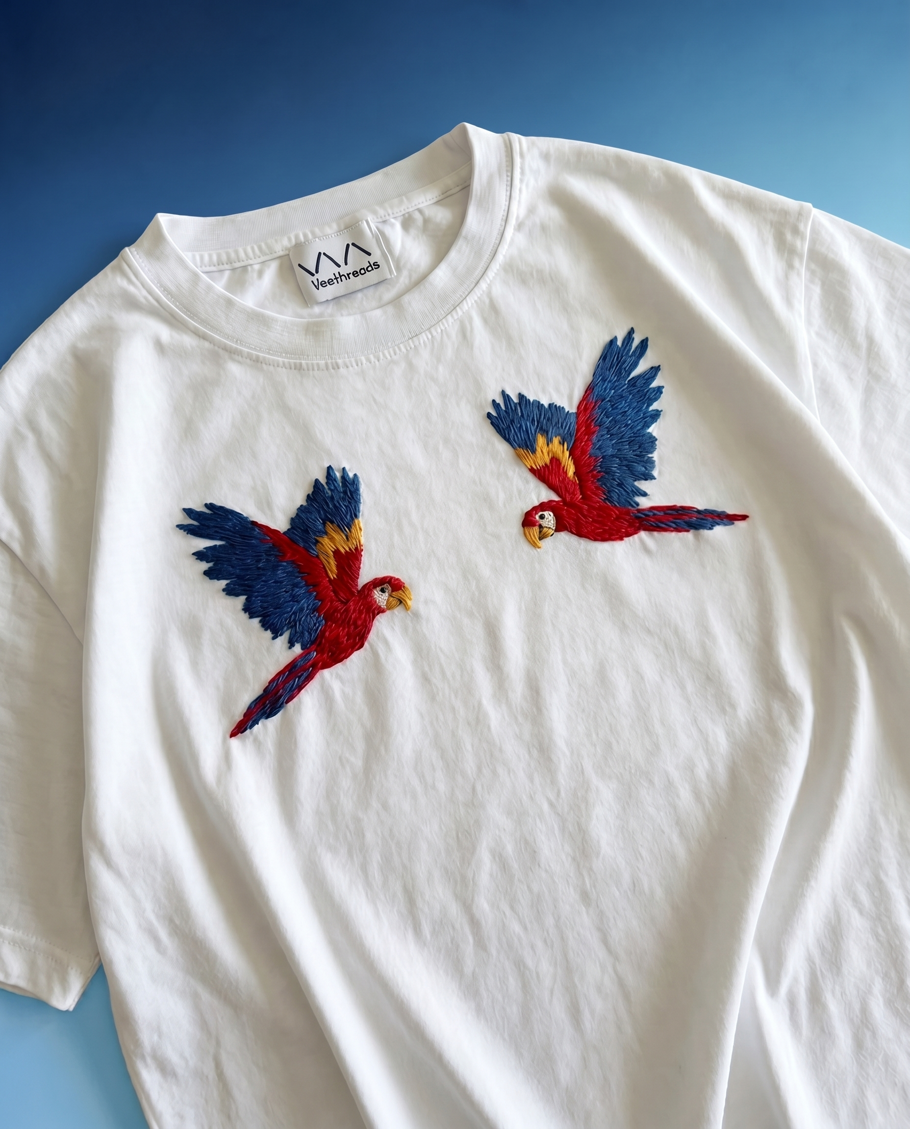 White t-shirt with embroidered parrot designs on a blue background