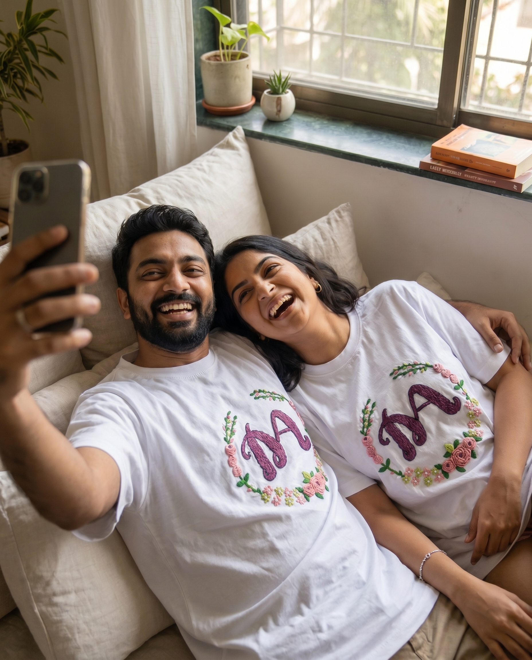 Man and woman sitting on a couch taking a selfie wearing matching name white t-shirts with decorative text.