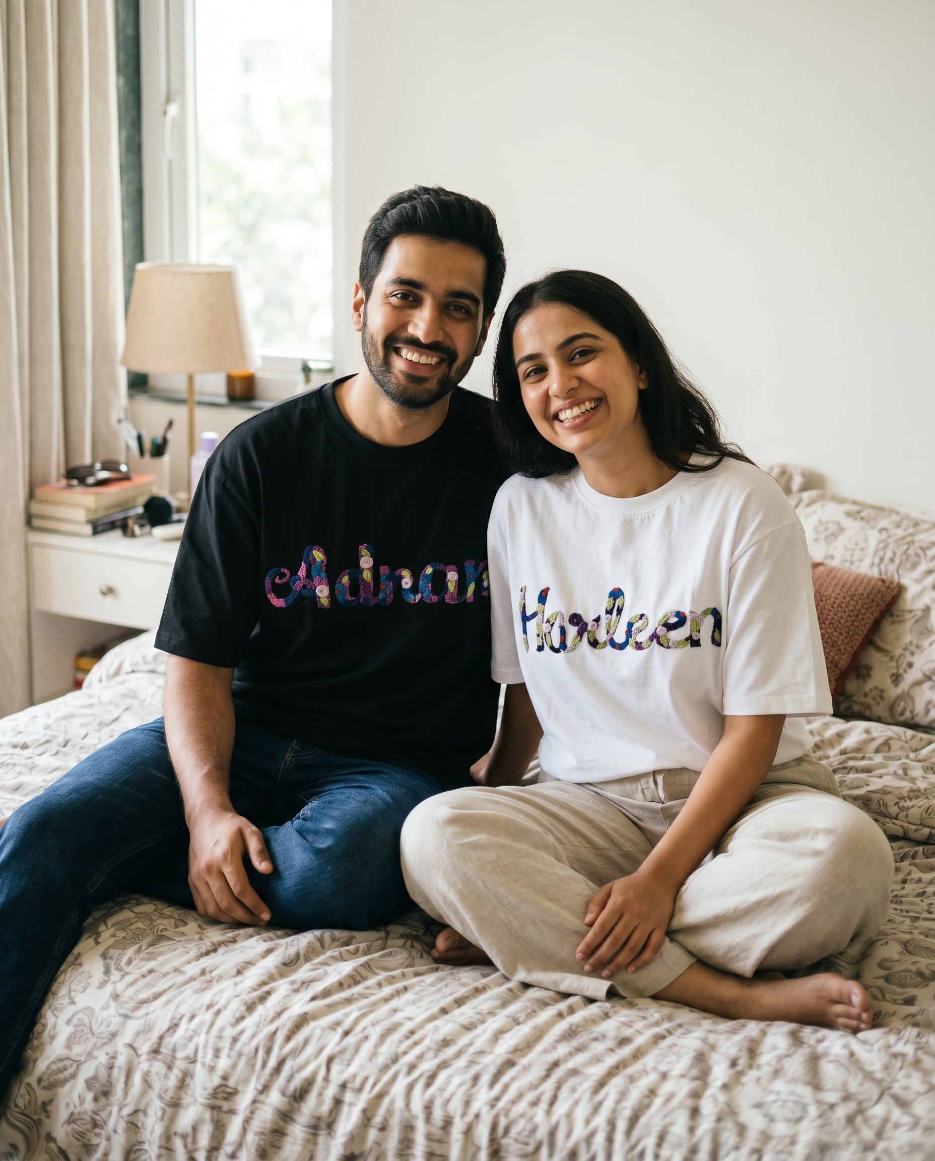 Man and woman sitting on a bed wearing customised name t-shirts with colorful text.