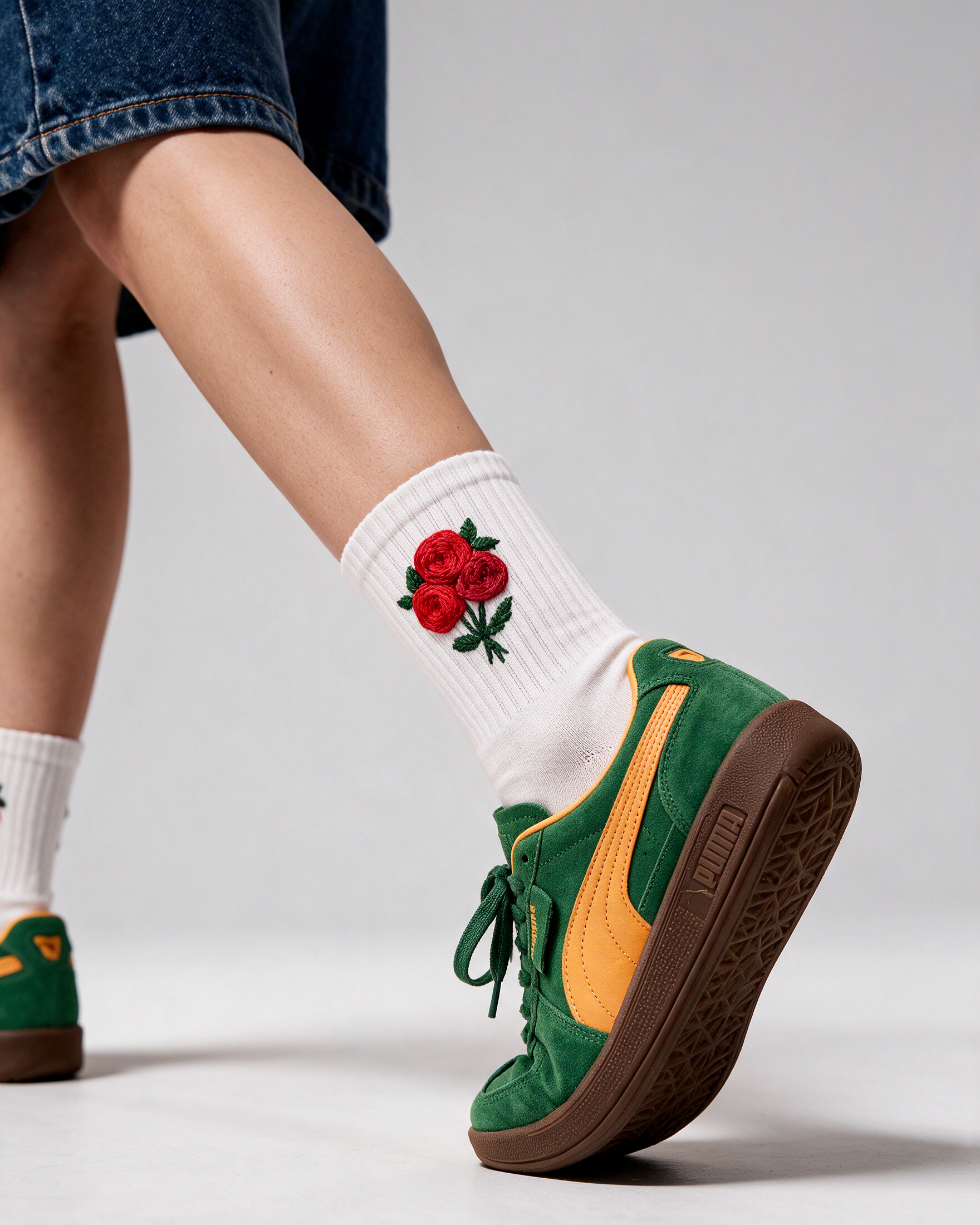 A person wearing green Puma sneakers with yellow and brown details, white crew socks featuring a red rose embroidery, and blue denim shorts, captured from a low angle against a light gray studio background.