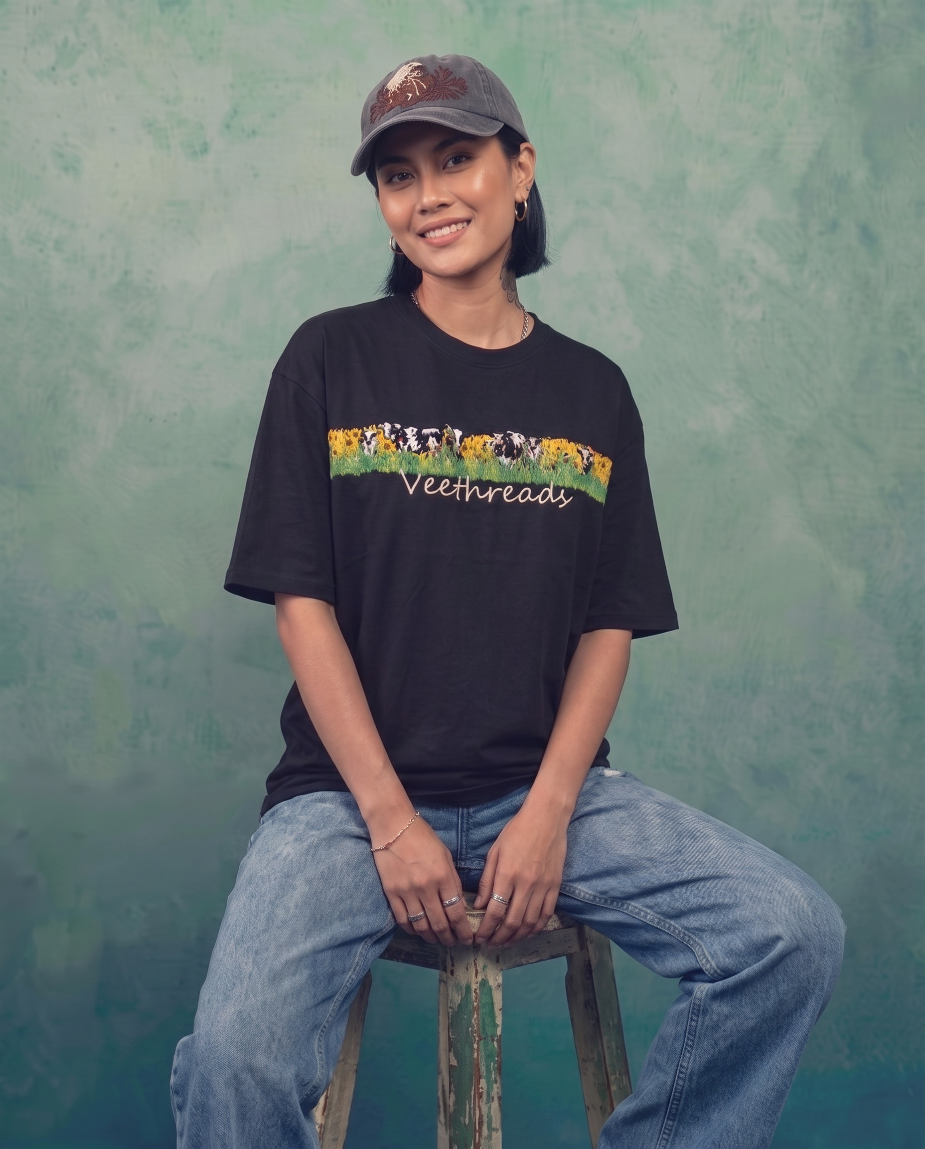 Woman seated wearing black hand embroidered oversized t-shirt with sunflower meadow and cows across the chest