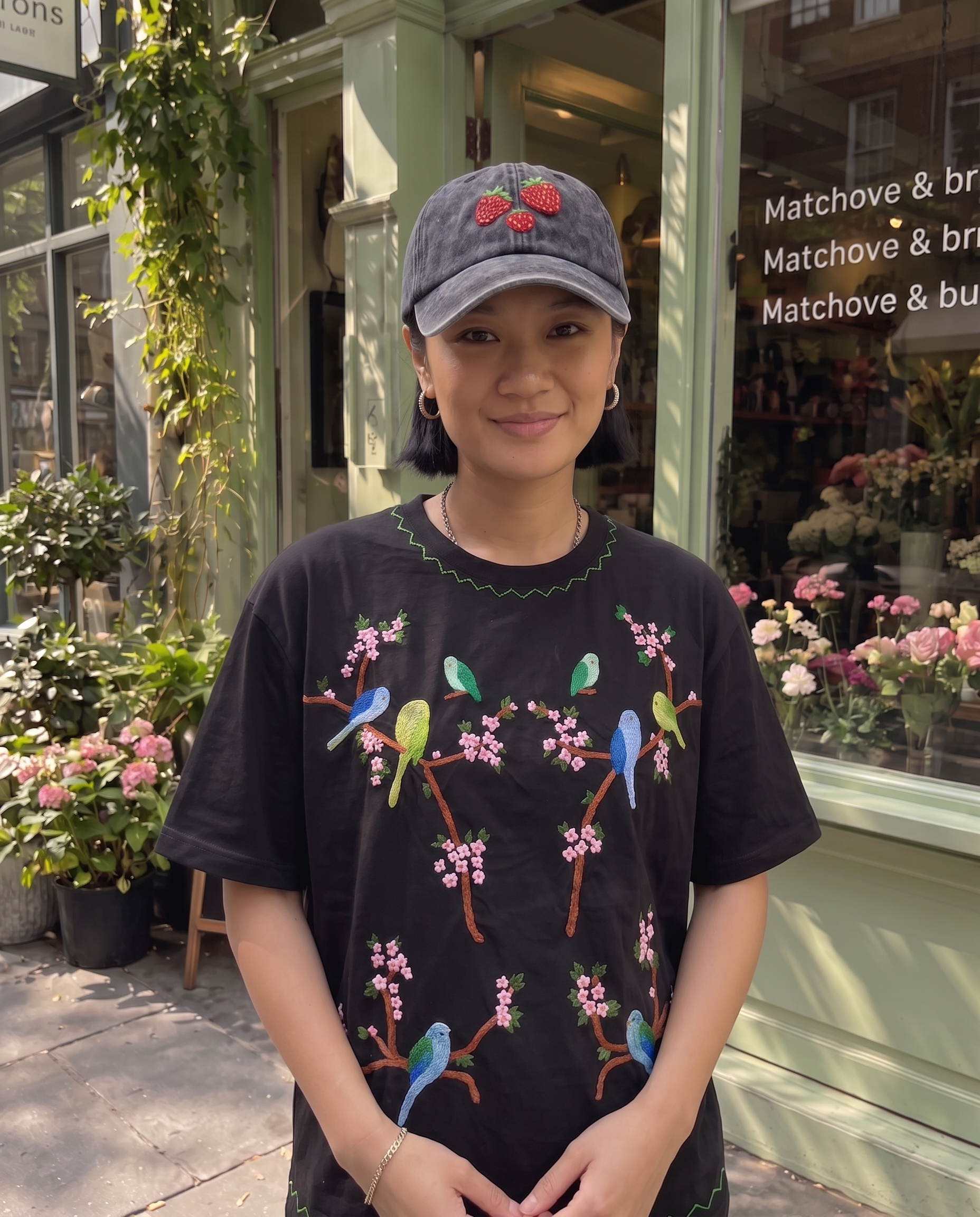 Woman in a washed black strawberry cap and embroidered parakeet tee outside a flower shop