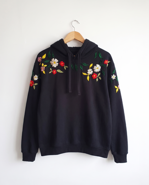 Black cotton fleece hoodie on a hanger with wildflowers hand-embroidered across the shoulders and sleeves