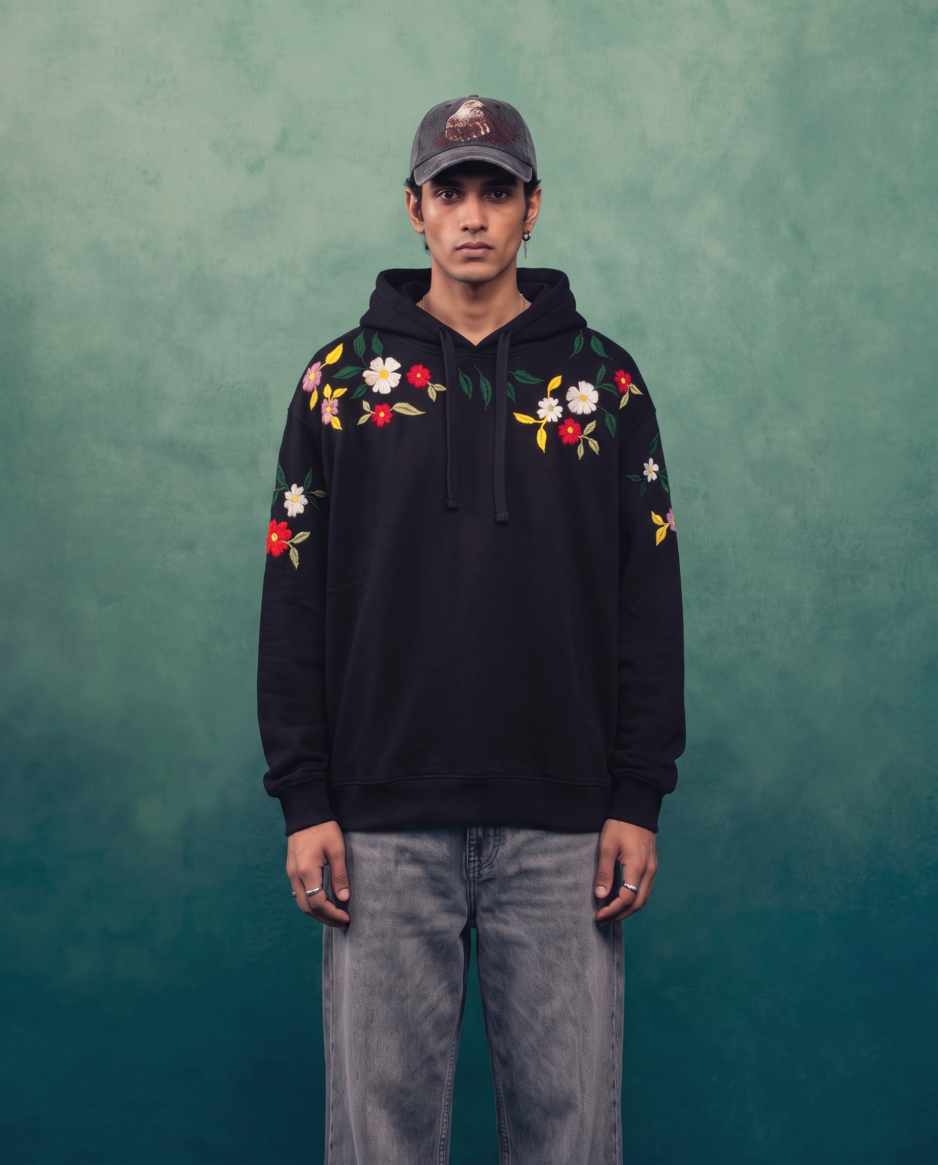 Man wearing the black floral hand-embroidered hoodie with jeans against a green backdrop