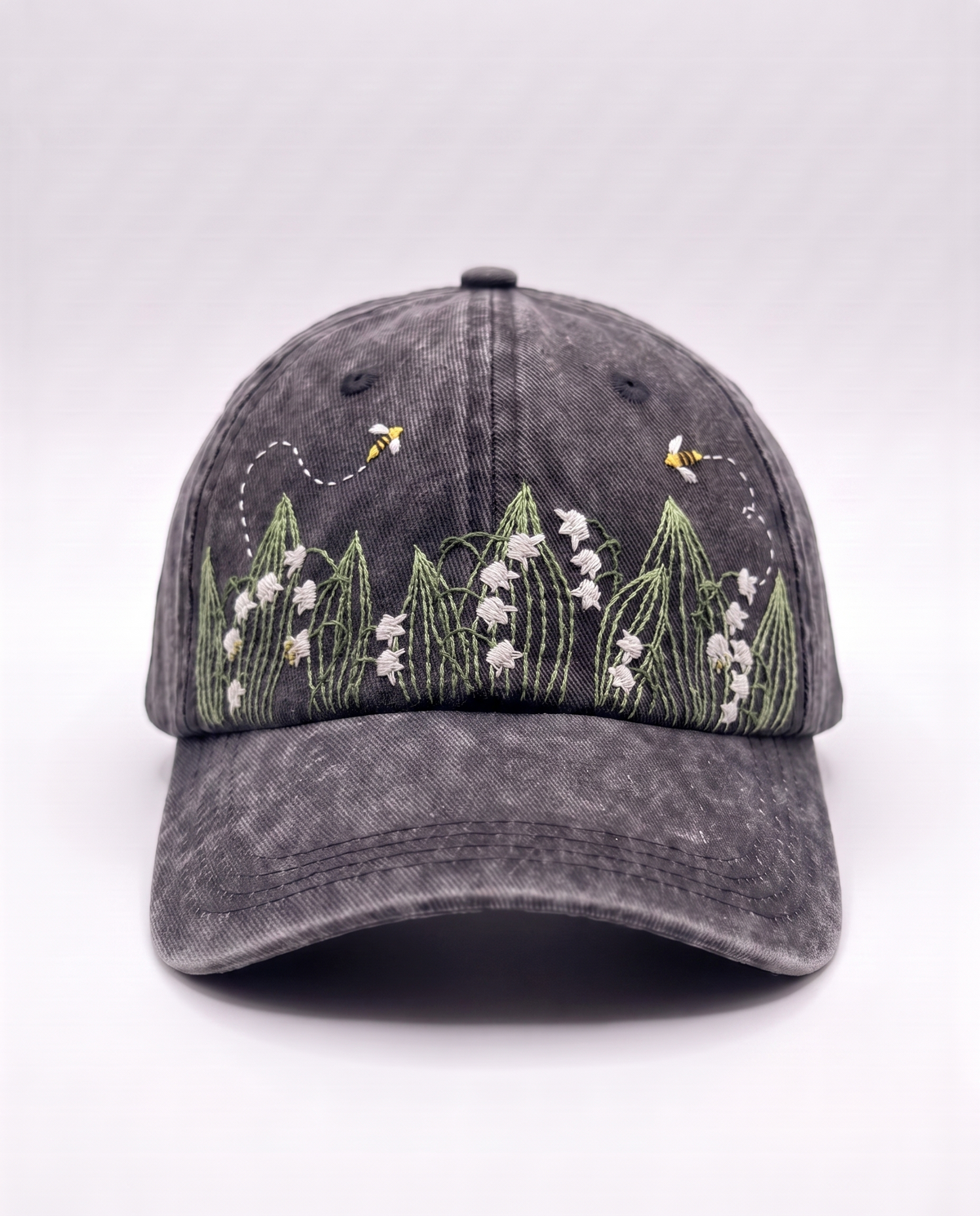 Charcoal acid-wash denim cap, front view, with white lily of the valley bells and two embroidered bees above them