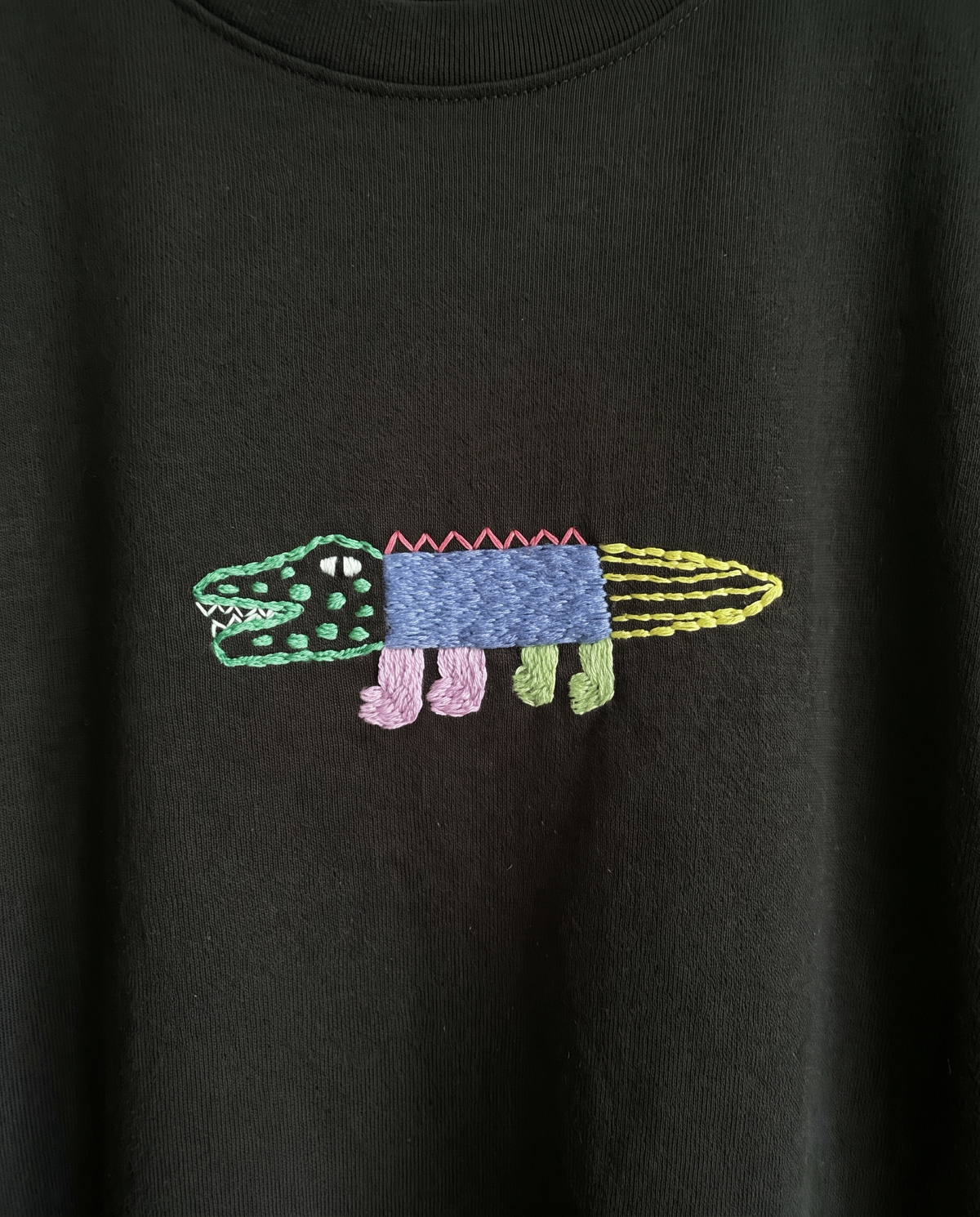 Hand-embroidered crocodile with a pink zigzag back and yellow tail on black cotton, straight-on view