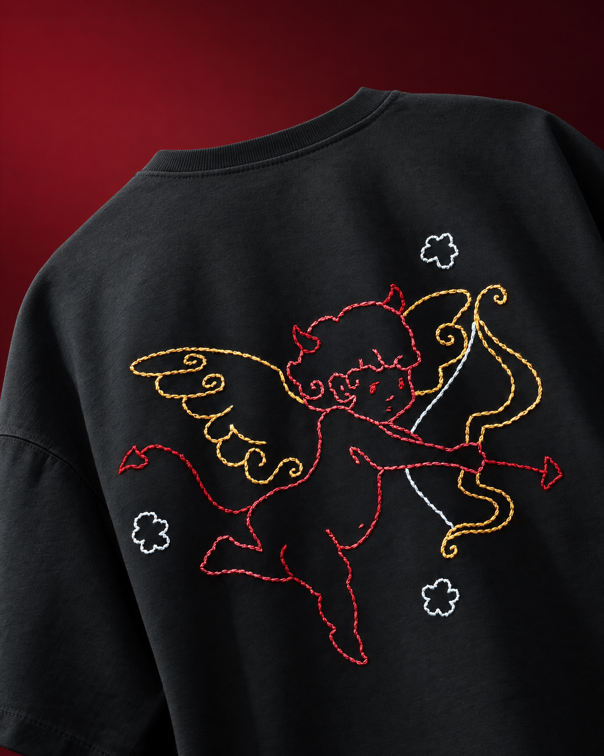 Close-up of the back of a black oversized T-shirt featuring a large embroidered cupid design in red, yellow, and white, with small white cloud motifs, set against a dark red background.